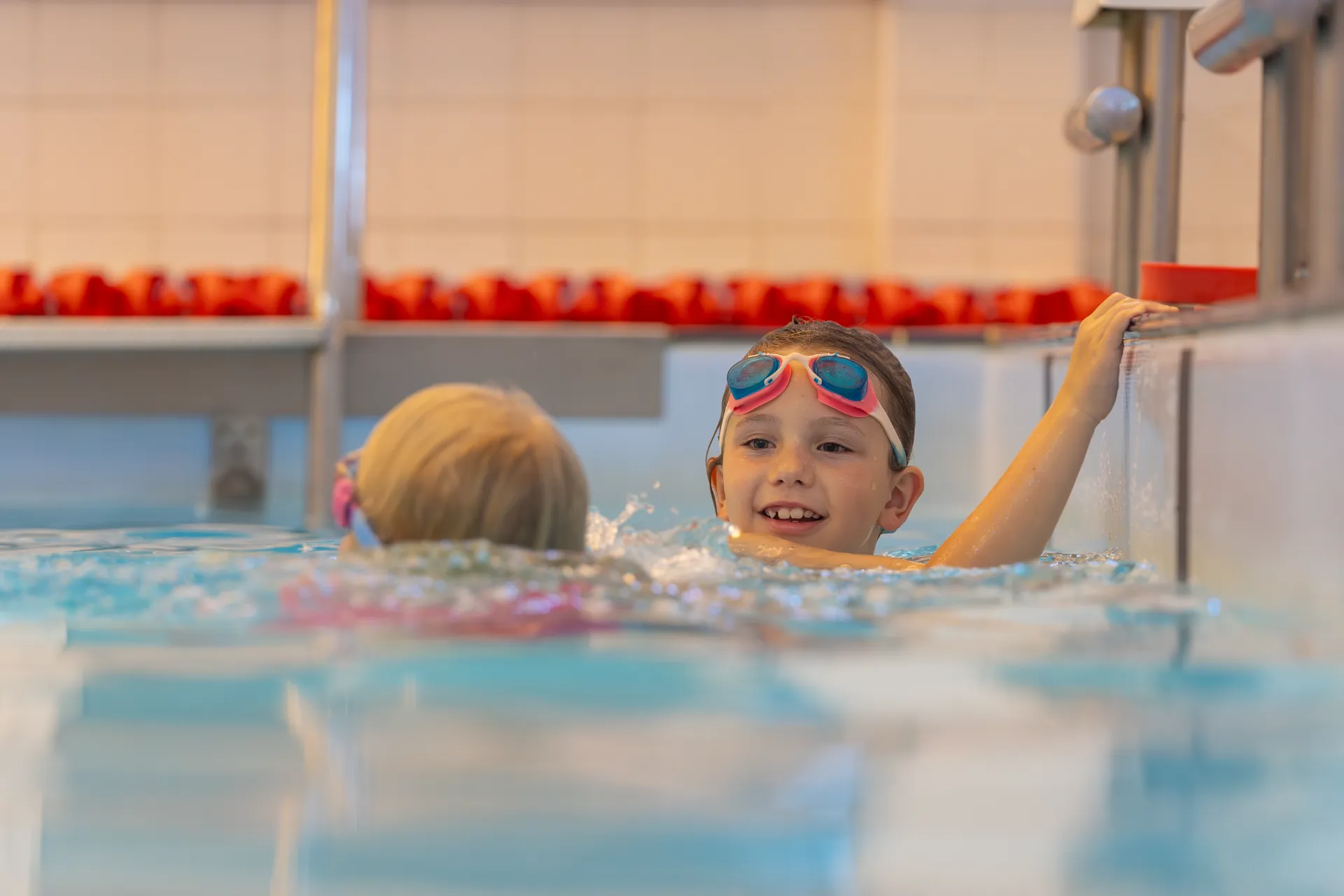 Smiling child holding on to the side of a swimming pool while talking to her friend